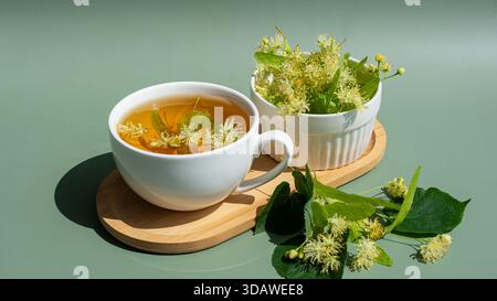 Una rilassante tazza di tiglio alle erbe con fiori freschi di tiglio esposti in una tazza bianca. Lo sfondo presenta una superficie verde rilassante che migliora il Foto Stock