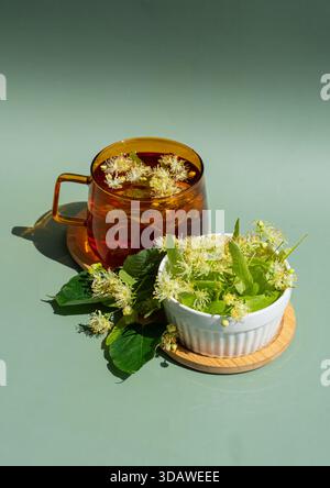 Dall'alto, una tazza di vetro piena di tiglio alle erbe è circondata da fiori di tiglio freschi in un piatto bianco, il tutto su una superficie verde liscia, creat Foto Stock