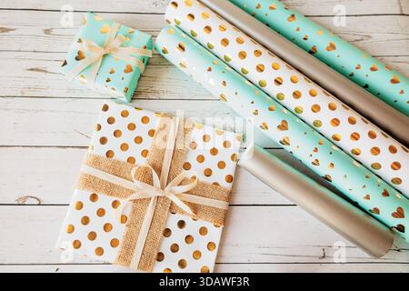 Elegante scenario di confezione regalo con carta a puntini dorata e motivo a cuore, nastri e fiocchi. Piatto su legno vintage, perfetto per il marketing stagionale e. Foto Stock