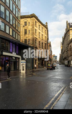 Glasgow, Regno Unito - 22 novembre 2025: Vista di edifici in arenaria che si illuminano sotto un cielo morbido, riflesso nelle strade bagnate, come un solitario pedone Foto Stock