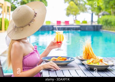 Donna al sole che beve un drink tropicale a bordo piscina, indossando l'estate Foto Stock