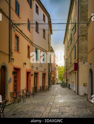 Stradina stretta e affascinante nel villaggio di Castagneto Carducci, Toscana. Vecchi edifici e vicoli lastricati di pietra con tavoli da caffe' all'aperto sotto un morbido cielo al tramonto Foto Stock