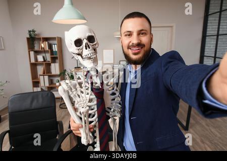 Giovane uomo d'affari con scheletro umano che fa selfie in ufficio Foto Stock