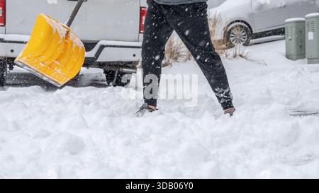 Individual sta spalando neve pesante da un vialetto con una pala da neve gialla, in mezzo a una scena invernale con fiocchi di neve che cadono intorno, sottolineando l'inverno Foto Stock