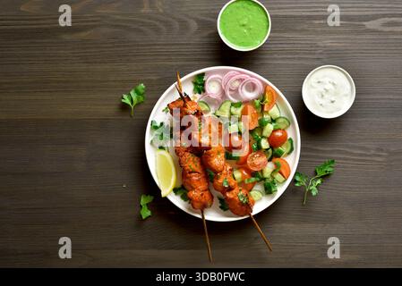 Gli spiedini Tandoori sono marinati in spezie, yogurt e succo di limone serviti con insalata di pomodoro e cetrioli su fondo rustico in legno. Vista dall'alto, disposizione piatta Foto Stock