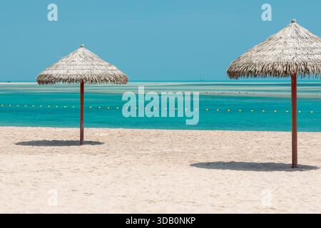 Tranquilla spiaggia con due ombrelloni di paglia su sabbia pallida, calme acque turchesi e un lontano orizzonte blu. Ideale per viaggi, vacanze e somme Foto Stock