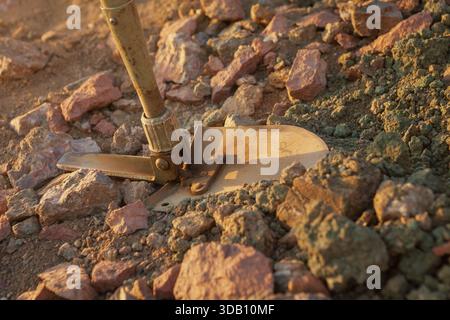 Una pala militare arrugginita scava nella terra rocciosa, rossastra-marrone, mostrando varietà geologiche, suggerendo scavi o esplorazioni. Foto Stock