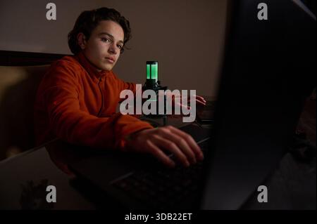 Un giovane con cappuccio arancione si siede a una scrivania con un notebook e un microfono verde brillante, focalizzato sullo schermo. Accogliente studio domestico per lo streaming e i gami Foto Stock
