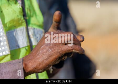 vista ravvicinata dei lavoratori africani delle mani, sporche e insensibili, dal lavoro manuale che indossa un giubbotto ad alta visibilità e una camicia a maniche lunghe, indumenti da lavoro di sicurezza Foto Stock