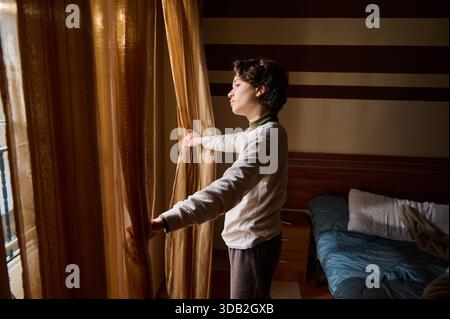 Una donna sta accanto a una grande finestra, tirando indietro tende dorate in una camera da letto con luce soffusa. Il momento trasmette calma, privacy e anticipazione di un nuovo da Foto Stock