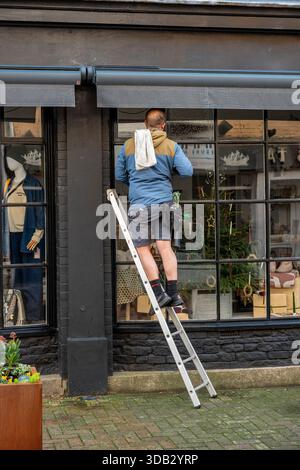 Detergente per vetri su scala posizionata in modo precario per raggiungere la parte superiore del finestrino, pulizia dei vetri su una scala, lavoro in altezza. Foto Stock