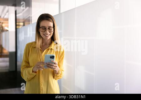 Giovane donna professionale elegante e positiva con occhiali che scrivono sullo smartphone Foto Stock