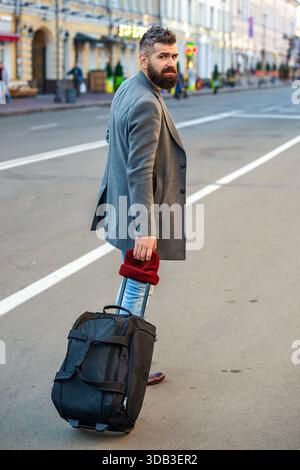 Un uomo d'affari in viaggio. Viaggiatore con barba che cammina lungo la strada della città. Turista con borsa in città. Barbuto uomo elegante che cammina e attraversa Foto Stock