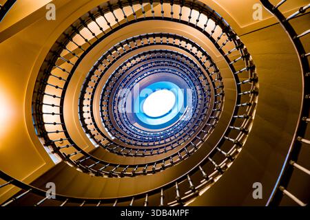 Guardando verso l'alto attraverso una scala circolare verso un lucernario ovale - balaustra in ferro concentrica e luce dorata calda Foto Stock