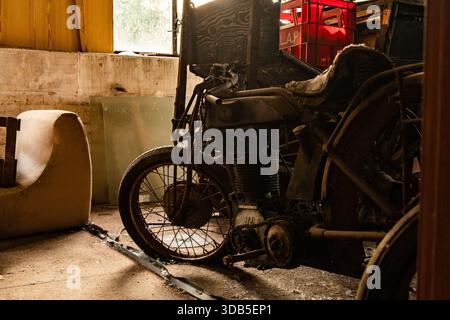 Motocicletta d'epoca polverosa con motore arrugginito in un'officina abbandonata illuminata da una luce soffusa sui finestrini Foto Stock