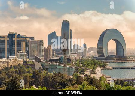 Baku, Azerbaigian - 10 maggio 2024: Paesaggio urbano caratterizzato dall'iconico Crescent Hotel, dalla SOCAR Tower e dall'Hilton hotel. Architettura moderna, parco verde e lungomare sotto un cielo nuvoloso Foto Stock