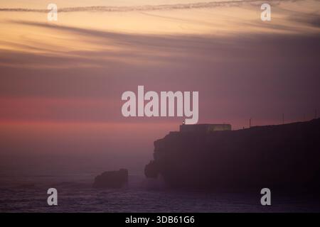 I colori tenui del tramonto si fondono con la nebbia costiera intorno alle scogliere di Nazaré, Portogallo. Il faro in cima al forte de São Miguel Arcanjo è visibile sulla t Foto Stock