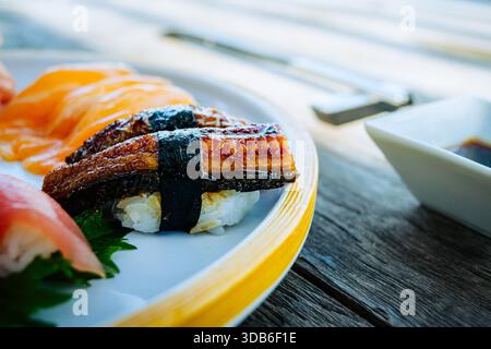 Il sushi Unagi Nigiri, caratterizzato da anguille cotte glassate con salsa, si appoggia accanto al nigiri di salmone su un piatto di ceramica accentuato da un bordo giallo. Chopstic in legno Foto Stock