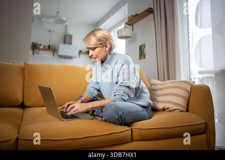 Donna d'affari di mezza età concentrata che lavora comodamente con un computer portatile su un comodo divano nel soggiorno. Foto Stock