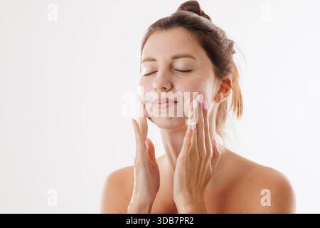 Giovane donna con una pelle luminosa che si lava il viso con un detergente delicato, godendo di una routine di cura della pelle, isolata su sfondo bianco. Foto Stock