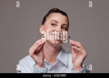Giovane donna sorridente con bretelle che reggono occhiali rotondi, espressione sicura e felice, elegante in una camicia bianca, ritratto moderno su sfondo grigio. Foto Stock