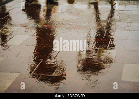 Sagome di persone che camminano con ombrelli riflessi sul marciapiede bagnato durante il giorno di pioggia nella città moderna. Concetto di stagioni, stile di vita urbano, Wea piovoso Foto Stock