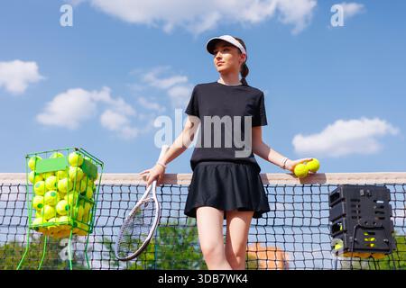 Giovane tennista sicura di sé che si appoggia su una rete con palle e racchetta sotto il cielo azzurro nelle giornate di sole. Foto Stock