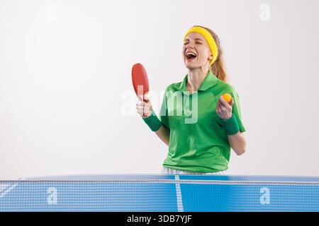 Una tennistavola eccitata con maglia verde e fascia gialla celebra la vittoria tenendo la palla e la pagaia. Foto Stock
