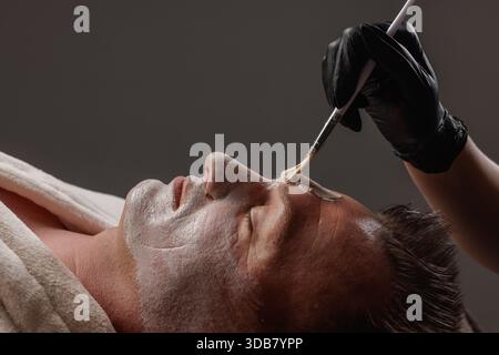 Uomo che riceve la maschera facciale con spazzola durante il trattamento di cura della pelle nel centro benessere, vista ravvicinata del relax e della cura del corpo. Foto Stock