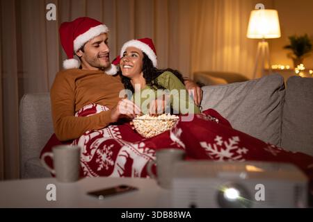 La coppia si diverte con un'accogliente serata di film natalizi nei cappelli di Babbo Natale mentre è avvolta in coperta a casa Foto Stock