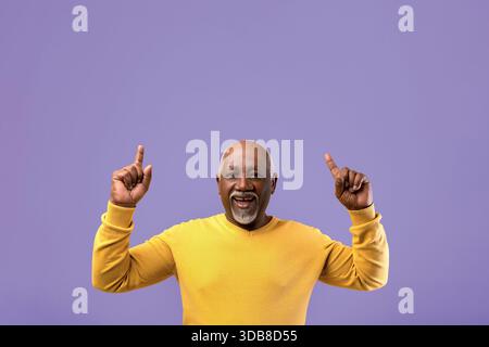 Uomo in maglione giallo alza entrambe le mani sorridendo su uno sfondo viola Foto Stock