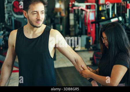 Personal trainer che assiste un cliente maschile con un braccio allungato o un esercizio fisico, concentrandosi sulla tecnica e la guida all'interno di un ambiente di palestra fitness per partecipare Foto Stock