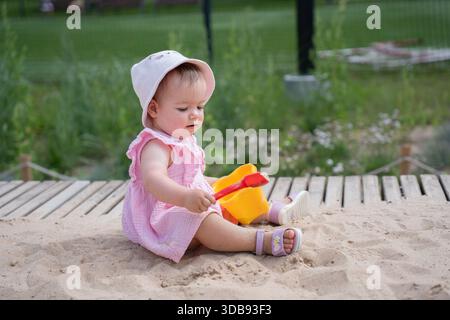 adorabile bambino che gioca con giocattoli di plastica colorati in una calda giornata di sole nella sandbox, circondato da vegetazione lussureggiante e tavole di legno in un parco all'aperto Foto Stock