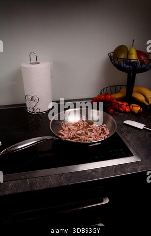 cucina con pancetta frizzante in una padella su un piano cottura moderno accanto a cesto di frutta fresca e asciugamani di carta, creando un'atmosfera di cucina vivace, fresca, Foto Stock