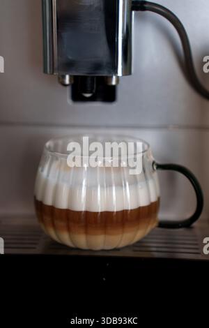 caffè a strati in una tazza di vetro decorata sotto una macchina da caffè metallica con manico scuro, con superficie cremosa e ricchi strati marroni in un set da caffè Foto Stock