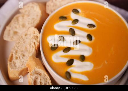zuppa cremosa di zucca adornata con vortici di yogurt e semi di zucca, accompagnata da fette di baguette croccante appena sfornata per un pasto caldo autunnale, Foto Stock