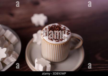 ambiente accogliente di una cremosa cioccolata calda con spruzzi di cioccolato in una tazza testurizzata su un tavolo di legno, circondato da marshmallow a forma di stella che evocano un wa Foto Stock