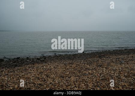 Tranquilla scena costiera, ciottoli grigi incontrano l'oceano calmo sotto cieli ricoperti, tranquilli e naturali. Foto Stock
