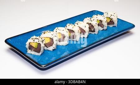 Involtini di sushi uramaki di tonno e avocado con semi di sesamo serviti su un piatto di ceramica blu isolato su sfondo bianco. Foto Stock