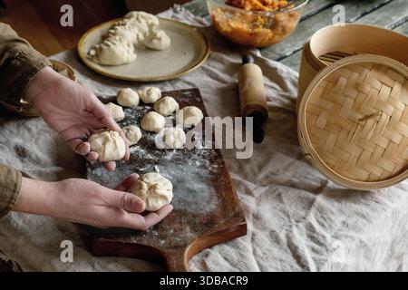 Preparazione di gnocchi asiatici al vapore con ripieno di verdure. Mani femminili che modellano gnocchi su tavola infarinata, vaporiera e ingredienti nelle vicinanze. Naturale Daylig Foto Stock