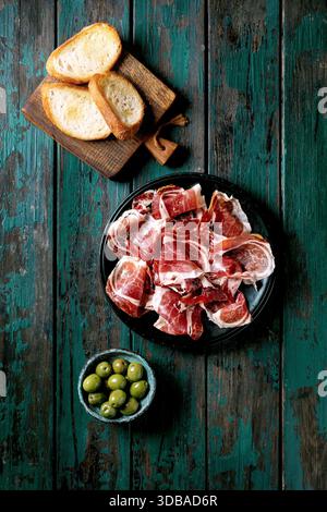 Prosciutto spagnolo a fette sottili Jamon Iberico su piatto nero. Ciotola di olive verdi, pane bianco tostato. Vecchio sfondo in legno, turchese scuro, vista dall'alto. Foto Stock