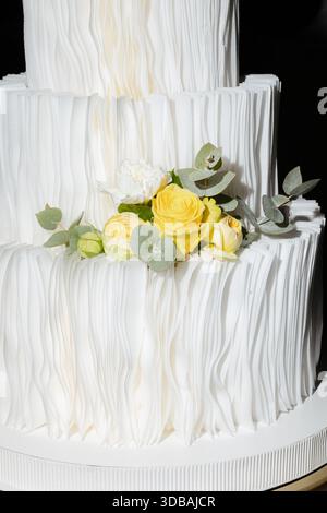 Elegante torta nuziale a tre piani con fondente bianco testurizzato e rose gialle fresche, perfetta come punto centrale per qualsiasi evento speciale. Foto Stock