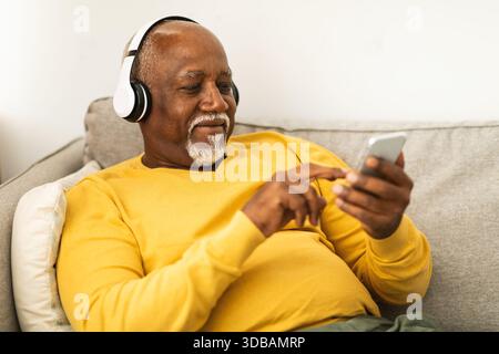 Un anziano sorridente ama ascoltare musica sullo smartphone mentre si rilassa sul divano nell'accogliente soggiorno del pomeriggio Foto Stock