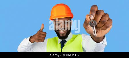 Il lavoratore mostra le chiavi con un sorriso su sfondo blu durante un progetto di costruzione Foto Stock