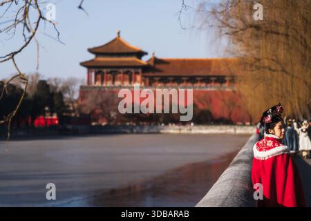 La vista di una donna in un vivace abbigliamento rosso tradizionale si erge lungo una sponda fredda del fiume con un suggestivo edificio rosso sullo sfondo, Pechino, Pechino, C. Foto Stock