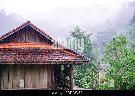 Tradizionale casa in legno con tetto piastrellato in un paesaggio montano nebbioso e piovoso, circondato da lussureggiante foresta tropicale nel sud-est asiatico. Foto Stock