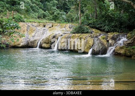 Le tranquille cascate si estendono su rocce coperte di muschio in una limpida piscina turchese, circondata da una lussureggiante giungla verde. Foto Stock