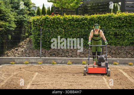 Un giardiniere lavora con una macchina per livellare e preparare il terreno in un giardino. L'ambiente presenta piante verdi e una recinzione. Foto Stock