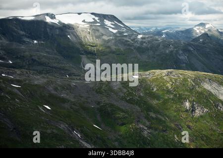 Le vette innevate si innalzano su paesaggi verdi con nuvole che si muovono attraverso il cielo nella regione montuosa della Norvegia. Foto Stock