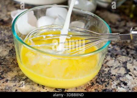 Il latte viene versato in un recipiente di vetro trasparente contenente uova sbattute, mescolando gli ingredienti umidi per ottenere una ricetta perfetta per impastare i waffle nella contea di Waukesha. Foto Stock